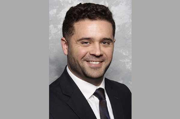 Professional headshot of a young man in a dark jacket and tie, posing with a friendly expression in front of a mottled background.