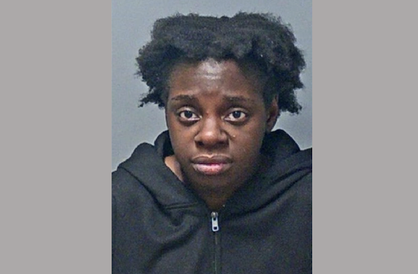 Booking-style portrait of a woman in a dark hooded sweatshirt with a neutral expression, centered in front of a gray backdrop.