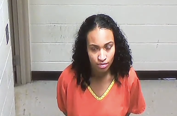 Woman in orange jail clothing with curly hair looks toward the camera in a booking photo.