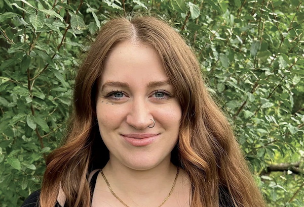 Young woman with wavy red hair and a small nose ring smiling outdoors with dense green foliage behind her.