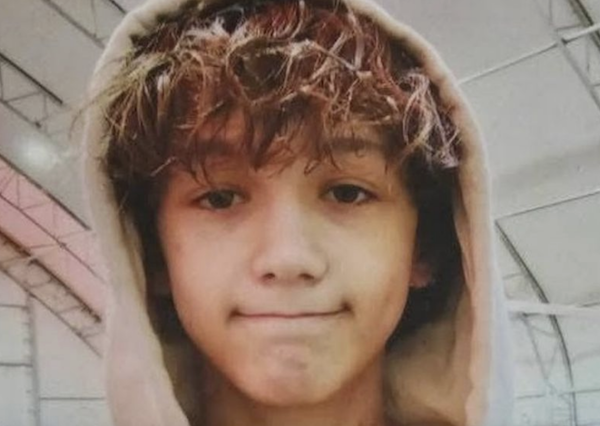 A child or young teen in a hoodie faces the camera with a slight closed-mouth expression in a softly lit indoor setting.