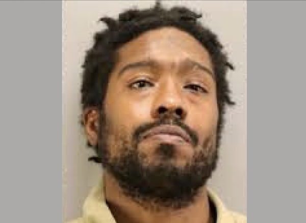 Head-and-shoulders booking photo of a man with facial hair and textured hair, wearing a light-colored shirt.