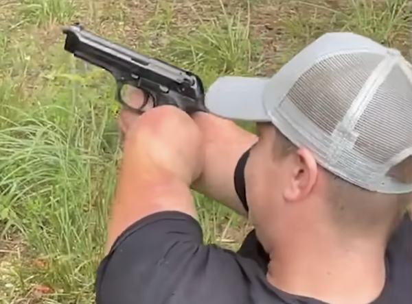 Outdoor photo of a person aiming a pistol to the left while turning their face toward the camera, standing beside tall grass and dirt ground.
