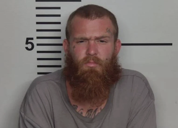 Head-and-shoulders image of a man with a large reddish-brown beard facing the camera against a marked wall.