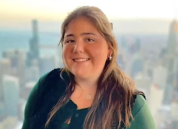 Portrait of a young woman in a dark green top, smiling at the camera with a high-rise city view behind her.