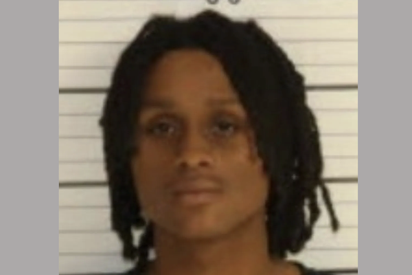 Front-facing booking photo of a person with dark dreadlocks and a neutral expression in front of measurement lines.