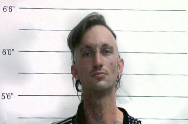 Mugshot-style image of a man with an undercut hairstyle, earrings, and neck tattoos standing in front of measurement lines.