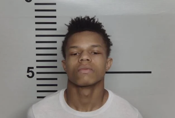 Portrait of a man with short textured hair and a neutral expression against a lineup wall marked with measurement lines.