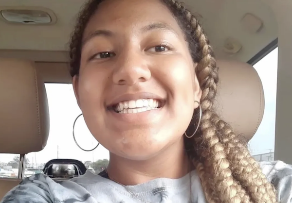 A young woman smiles at the camera from a vehicle with tan seats and daylight visible through the windows.