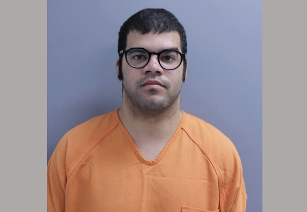 Front-facing mugshot of a man with short dark hair, glasses, and an orange detention shirt, standing against a solid neutral backdrop.