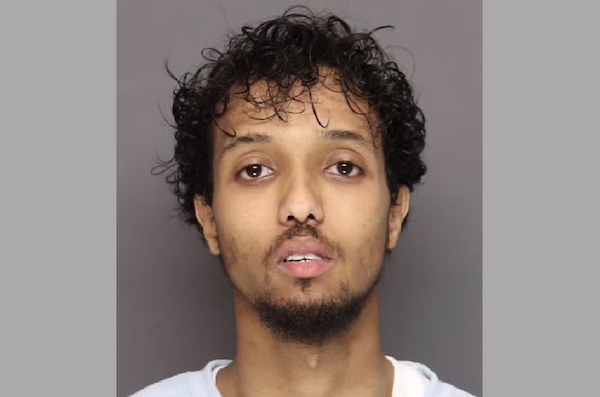 A young man in a light-colored shirt faces the camera in a tightly cropped portrait on a plain gray backdrop.