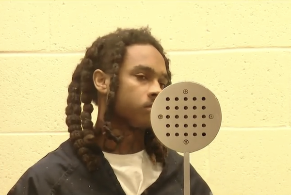 A man in dark clothing appears in a detention setting, partially obscured by a circular phone speaker.