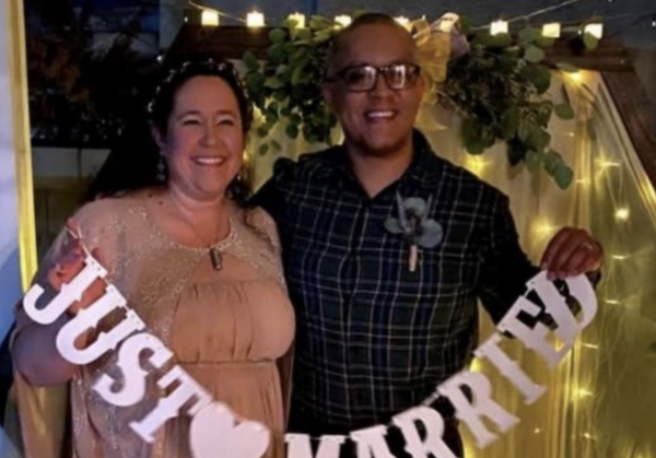 Wedding celebration photo of two people standing together in front of greenery and lights.