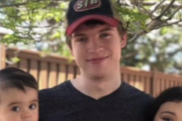 Young man wearing a baseball cap poses outdoors with a small child and a partially visible woman.