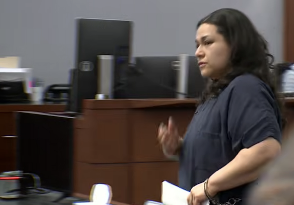 Courtroom image showing a person standing near counsel tables while holding papers.
