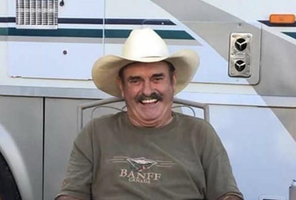 A man wearing a wide-brimmed cowboy hat laughs while seated outdoors beside an RV or trailer.
