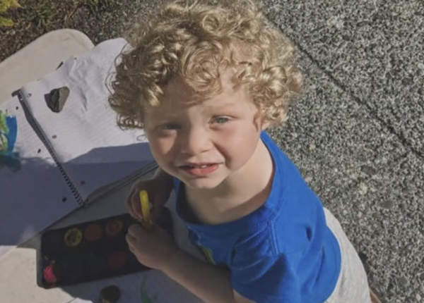 Child in a blue shirt looks up at the camera while painting on paper outdoors.