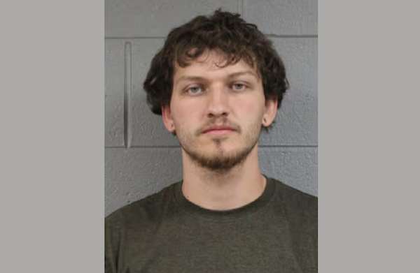 Mugshot-style portrait showing a young man with tousled hair, a short beard and a neutral expression.