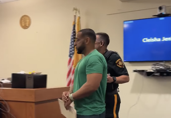 Side view of a handcuffed man walking past a courtroom bench with a sheriff’s deputy behind him.