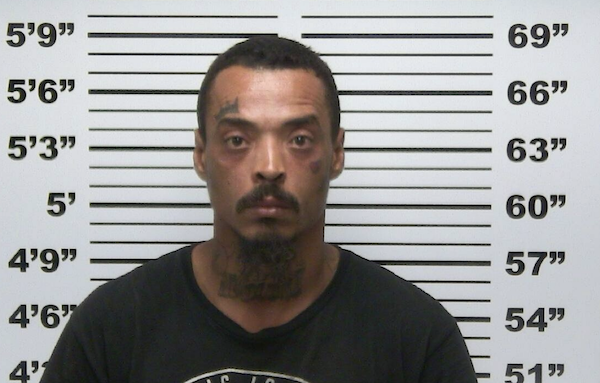 Front-facing mugshot of a man in a black shirt posed against a marked measurement backdrop.