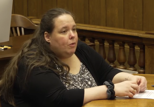 Side view of a seated woman addressing someone in a wood-paneled courtroom.
