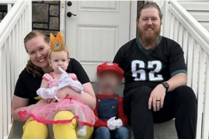 Smiling parents pose with two children in costumes outside a home; one child’s face is blurred.