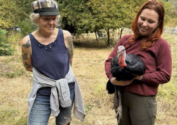 A red-haired woman cradles a black turkey as another woman wearing a cap stands beside her in a rural outdoor setting.