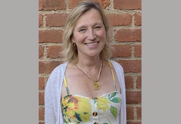 Portrait of a blonde woman smiling at the camera against a brick wall, dressed in a sunflower-patterned top with a white cardigan.