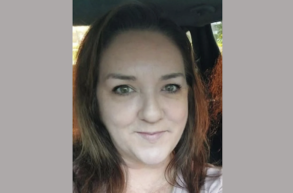 A woman with brown hair looks directly at the camera in a car, with daylight visible through the windows behind her.