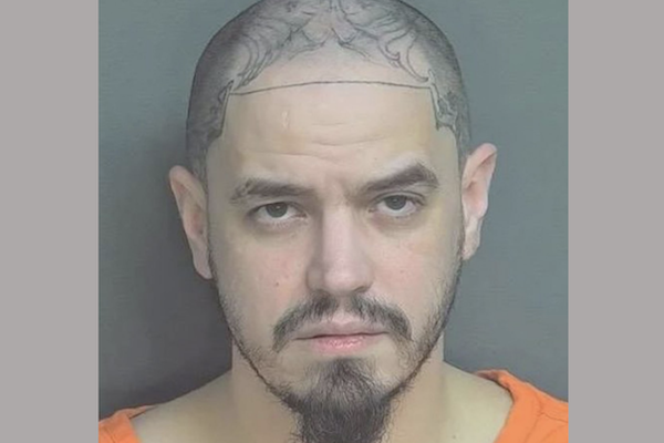 Front-facing mugshot of a man with extensive tattoos across his shaved scalp and dark facial hair against a gray background.