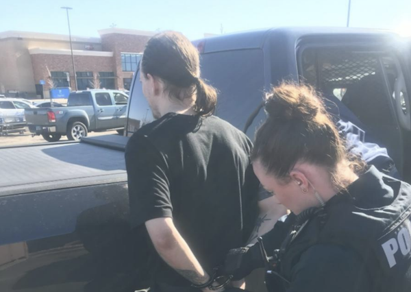 Outdoor scene showing a person near a dark vehicle as a police officer in tactical gear stands nearby.