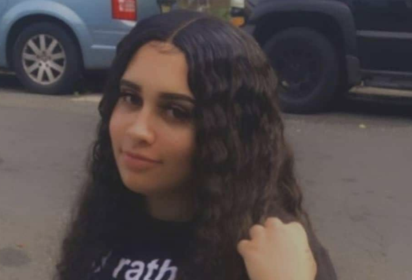 Close-up of a woman with long, wavy dark hair posing outside on a street with cars in the background.