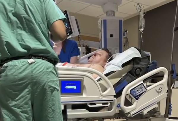 Hospital patient rests in an elevated bed as health care workers attend to them in a clinical treatment room.