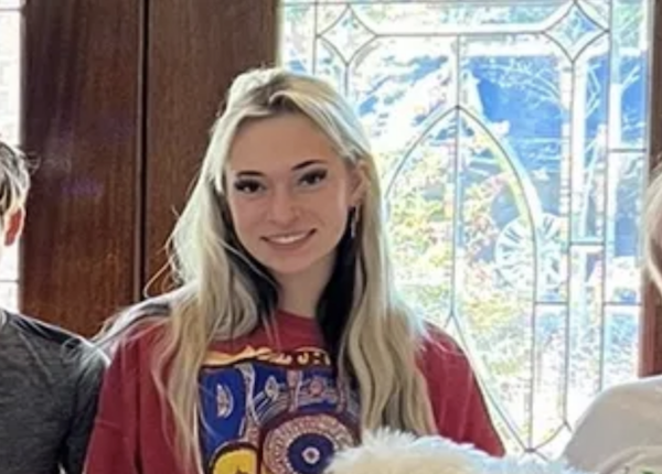 Blonde woman in a red graphic shirt smiles at the camera beside a bright patterned window.